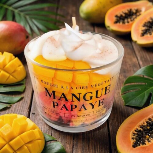 Bougie gourmande en verre montrant des couches de cire jaune (mangue) et rose (papaye) surmontées d'une chantilly de cire onctueuse et de morceaux de mangue trompe-l'œil, sur un fond de bois rustique et feuilles de palmier.