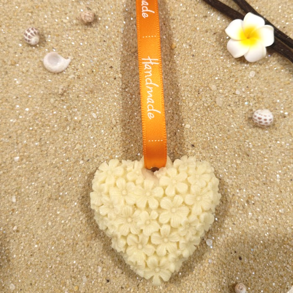 Suspension artisanale cĆur en cire vanille posĂ©e sur du sable fin avec gousses de vanille et coquillages.