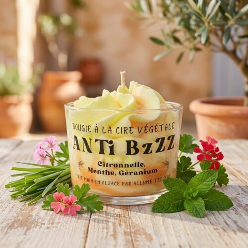 Bougie chantilly anti-moustique jaune et blanche Anti Bzzz d'Allume Tes Sens sur une table de terrasse en bois clair, entourée de brins de citronnelle, fleurs de géranium et feuilles de menthe.